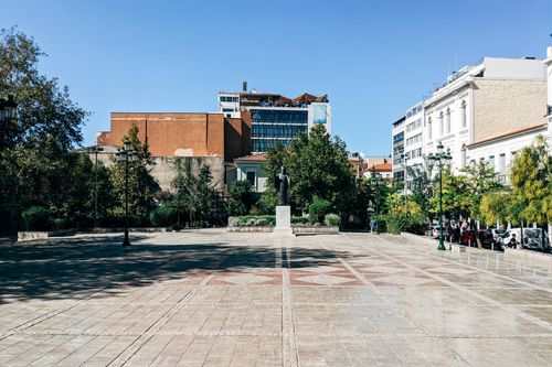 Free An open city plaza with a central statue surrounded by buildings and trees under a bright blue sky. Stock Photo