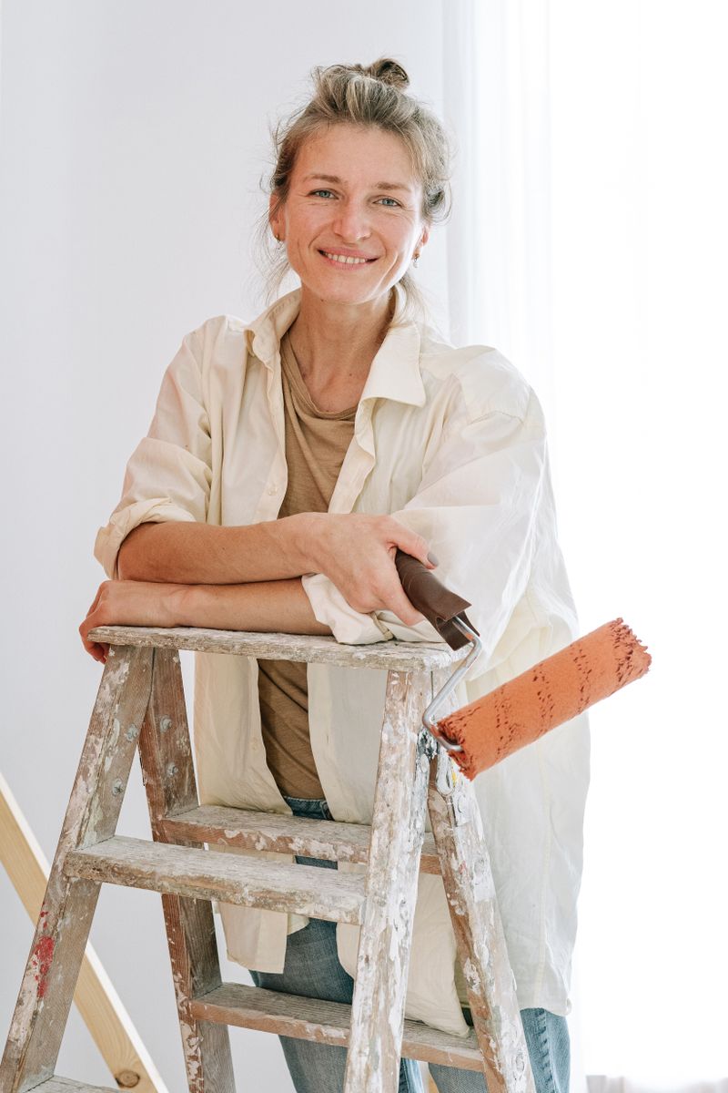 Maya Larkin, owner of Larkspur Painting Co., leaning on a paint-spattered ladder holding a roller