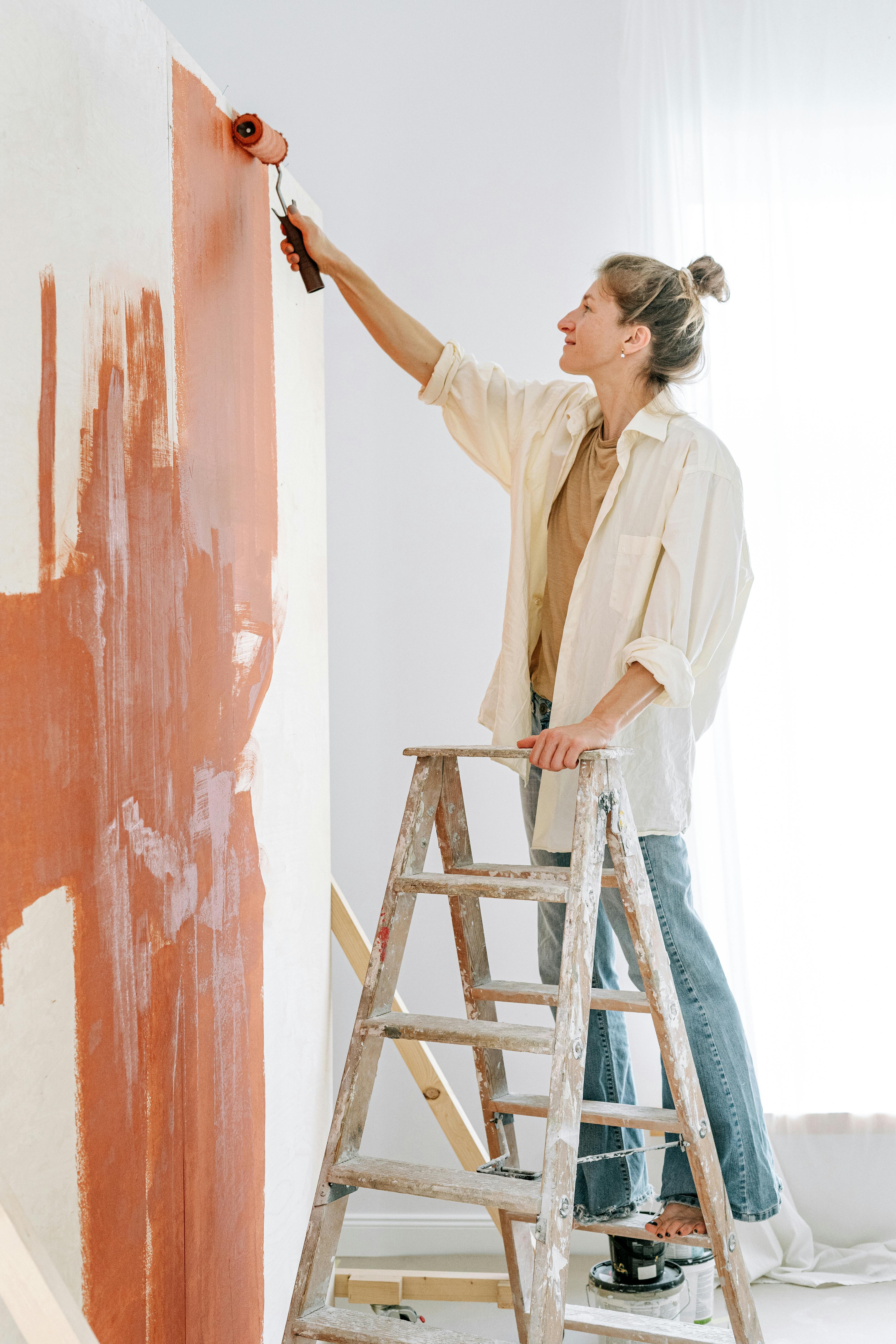 A woman painting a wall with a roller, standing on a ladder in a bright room.