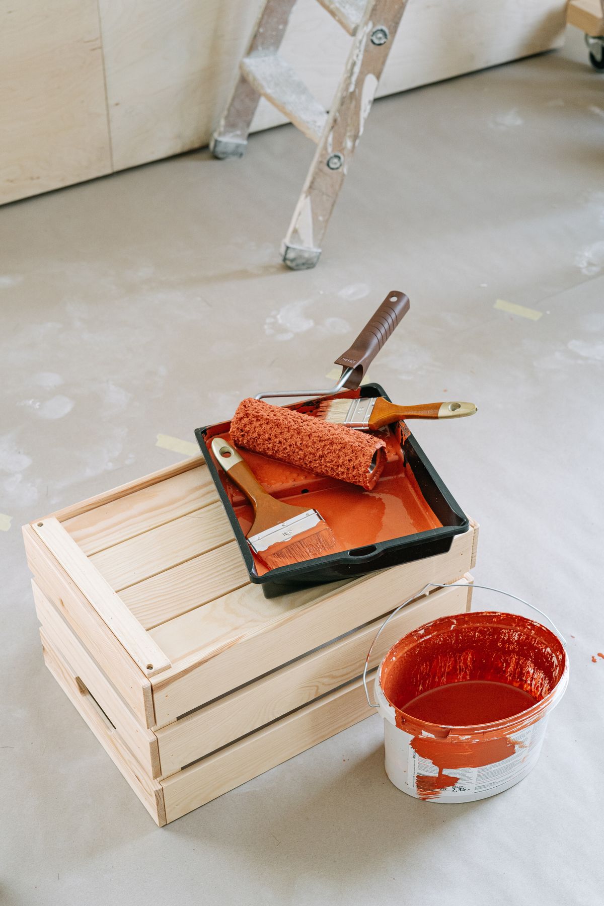A close-up of home renovation tools including paint roller and red paint bucket on a wooden crate. Illustrative editorial photograph.