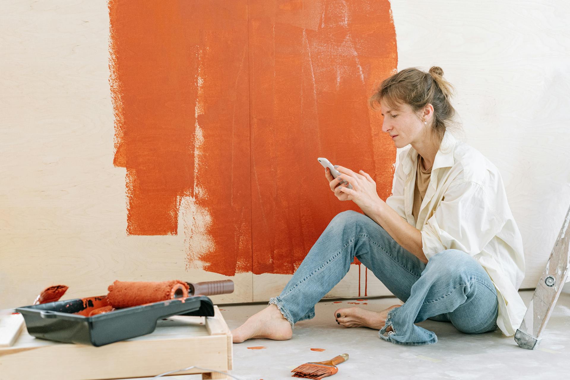 A woman takes a break from renovating by checking her phone while painting a wall.