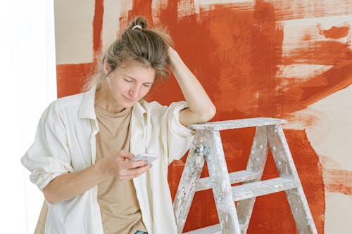 Woman in White Button Up Shirt Leaning on a Stepladder Using Cellphone