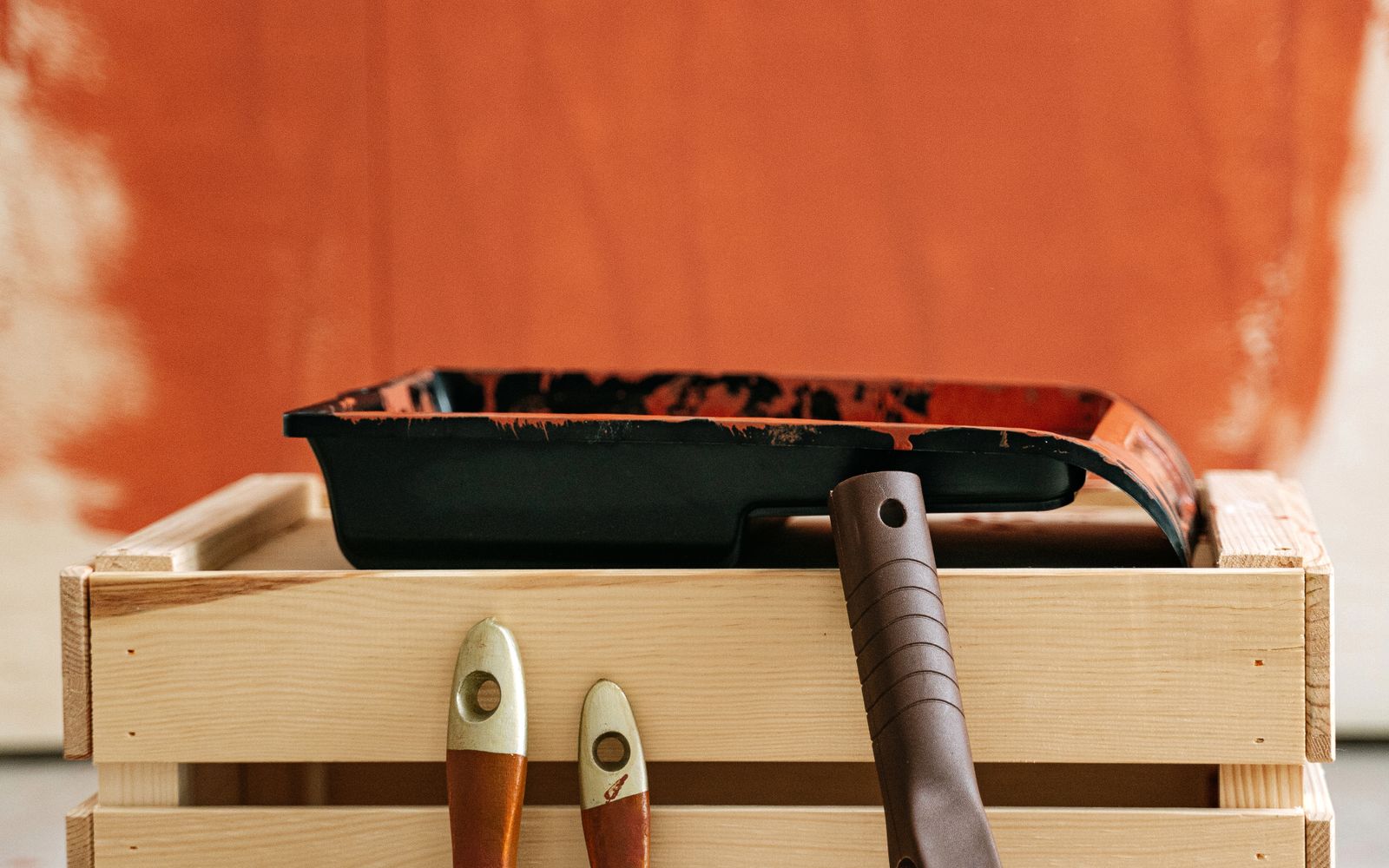 A wooden crate of brushes and a roller beside a partly painted wall