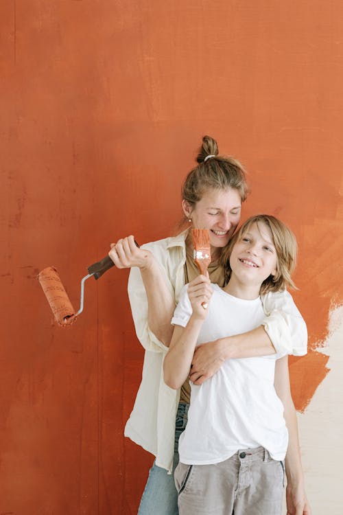 Woman in White Button Up Shirt Hugging a Boy Holding a Paintbrush