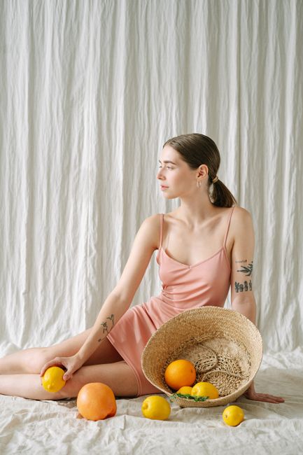 A woman in a pink dress sits with a basket of citrus fruits in a minimalist indoor setting.