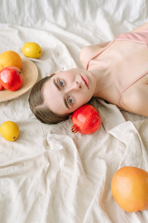 Woman Lying on White Textile Beside a Variety of Fruits