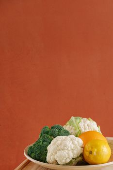 Plate of fresh broccoli, cauliflower, and citrus against a warm orange background, highlighting healthy food choices.