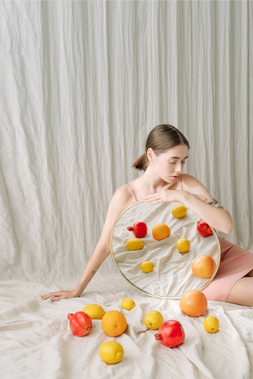 Woman in Pink Spaghetti Strap Dress Sitting on White Textile Holding a Mirror