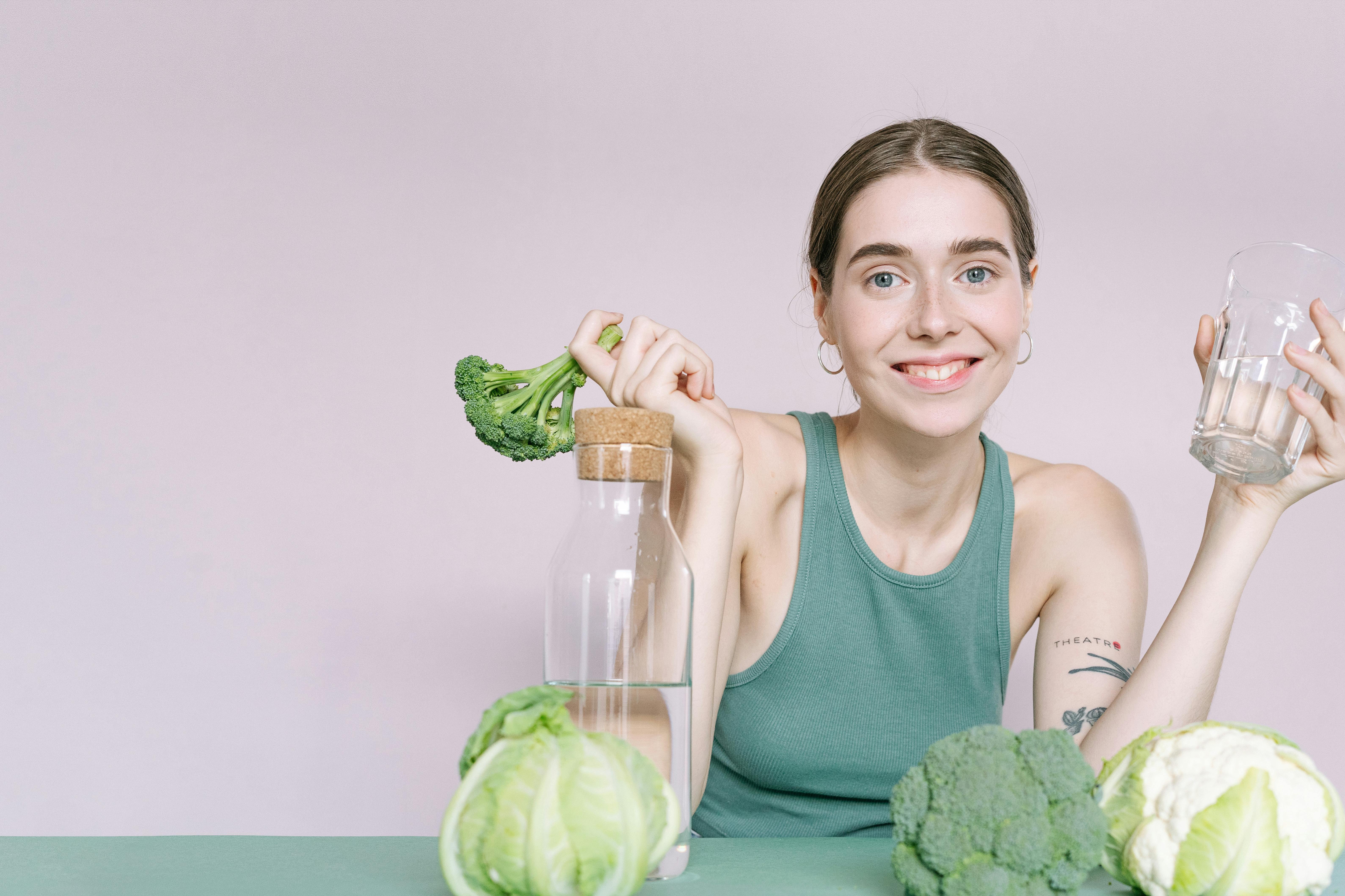 Free Woman in tank top smiling with broccoli, cabbage, and glass of water on table. Stock Photo