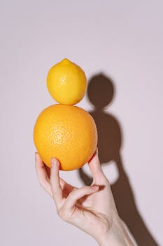 A hand balances an orange and a lemon, casting a creative shadow on a white background.
