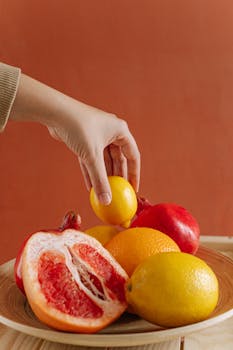 A close-up of a hand picking fresh citrus fruits from a wooden bowl against an orange background.