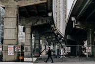 在现代化城市的市中心，一座旧立交桥下，一名身穿黑衣、身份不明的男子走在道路上，旁边的栅栏上挂着警告牌