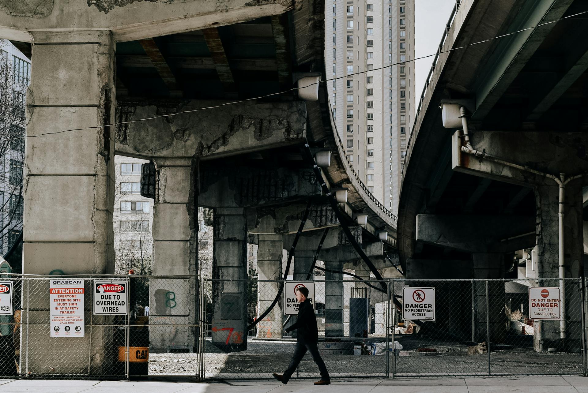 Unrecognizable male in black clothes walking down road with warning sign board on fence under old overpass in modern city downtown