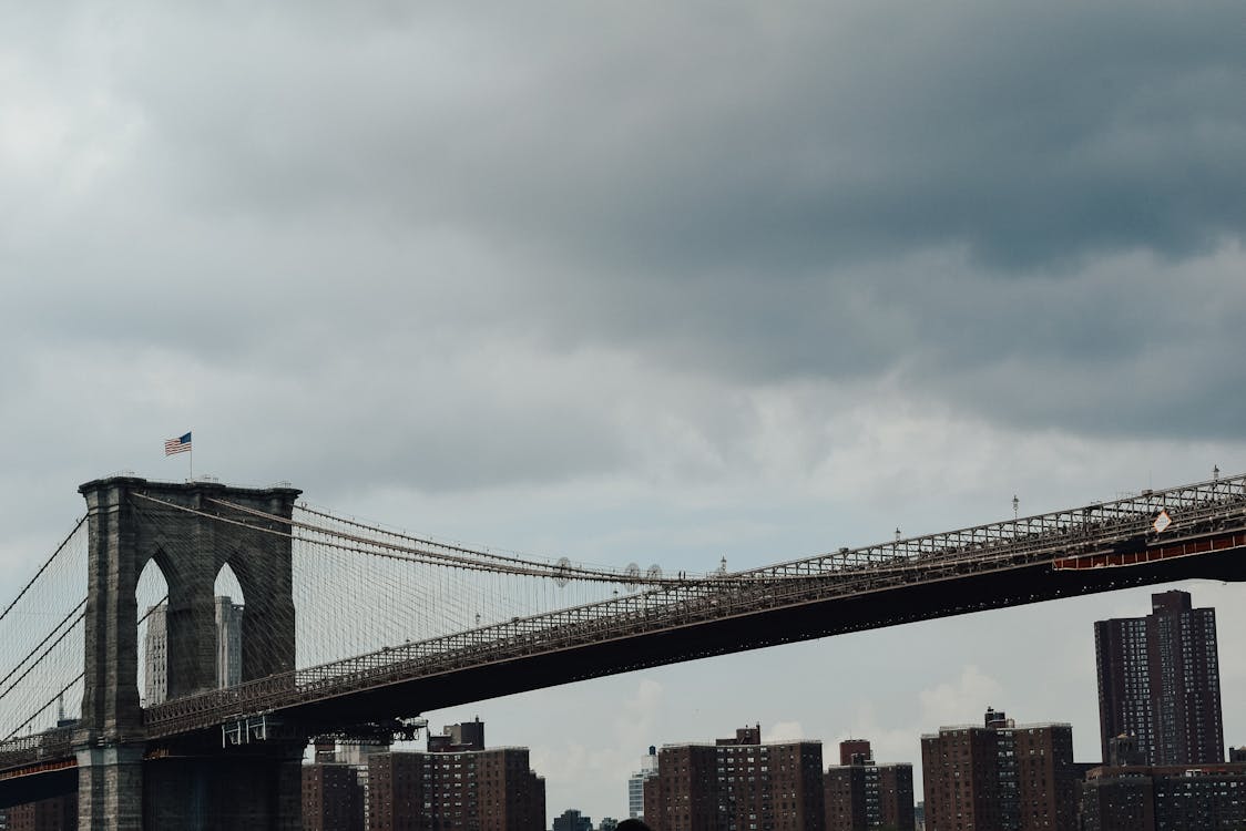 Low angle of Brooklyn bridge with waving American flag against high rise buildings and gloomy clouds