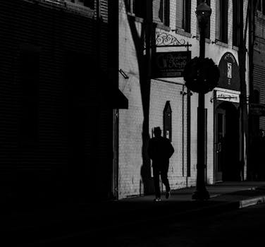Dramatic black and white photo of a man walking past buildings in a shadowy urban alleyway.