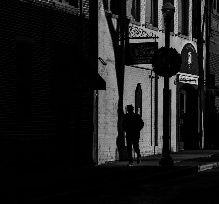 Dramatic black and white photo of a man walking past buildings in a shadowy urban alleyway.