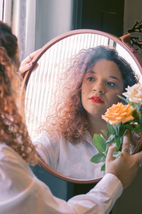 Free Back view of young female in white shirt with mirror in hands and flower looking at mirror in light room Stock Photo