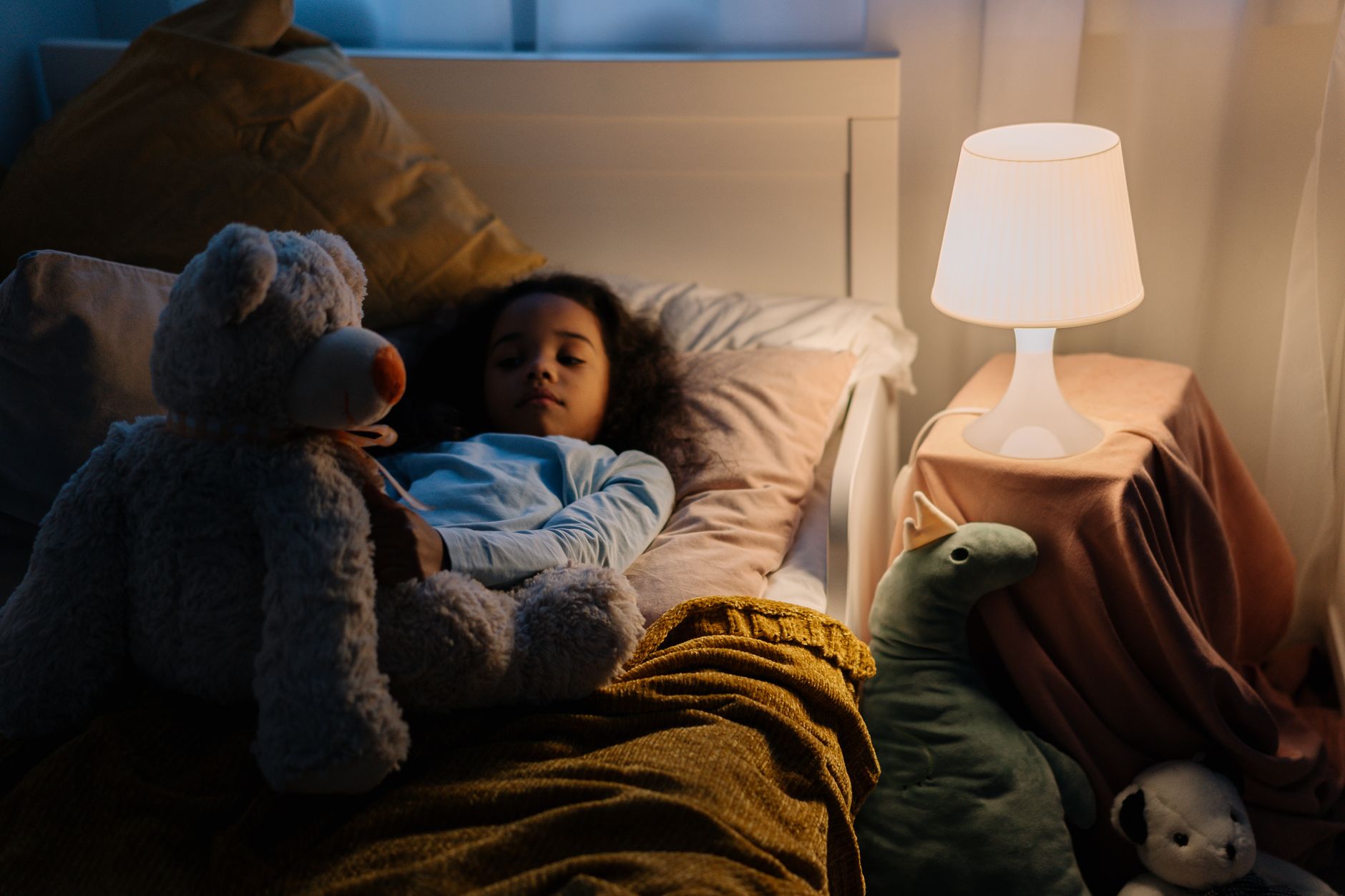 Young girl resting in bed with a teddy bear and toys, illuminated by a bedside lamp.