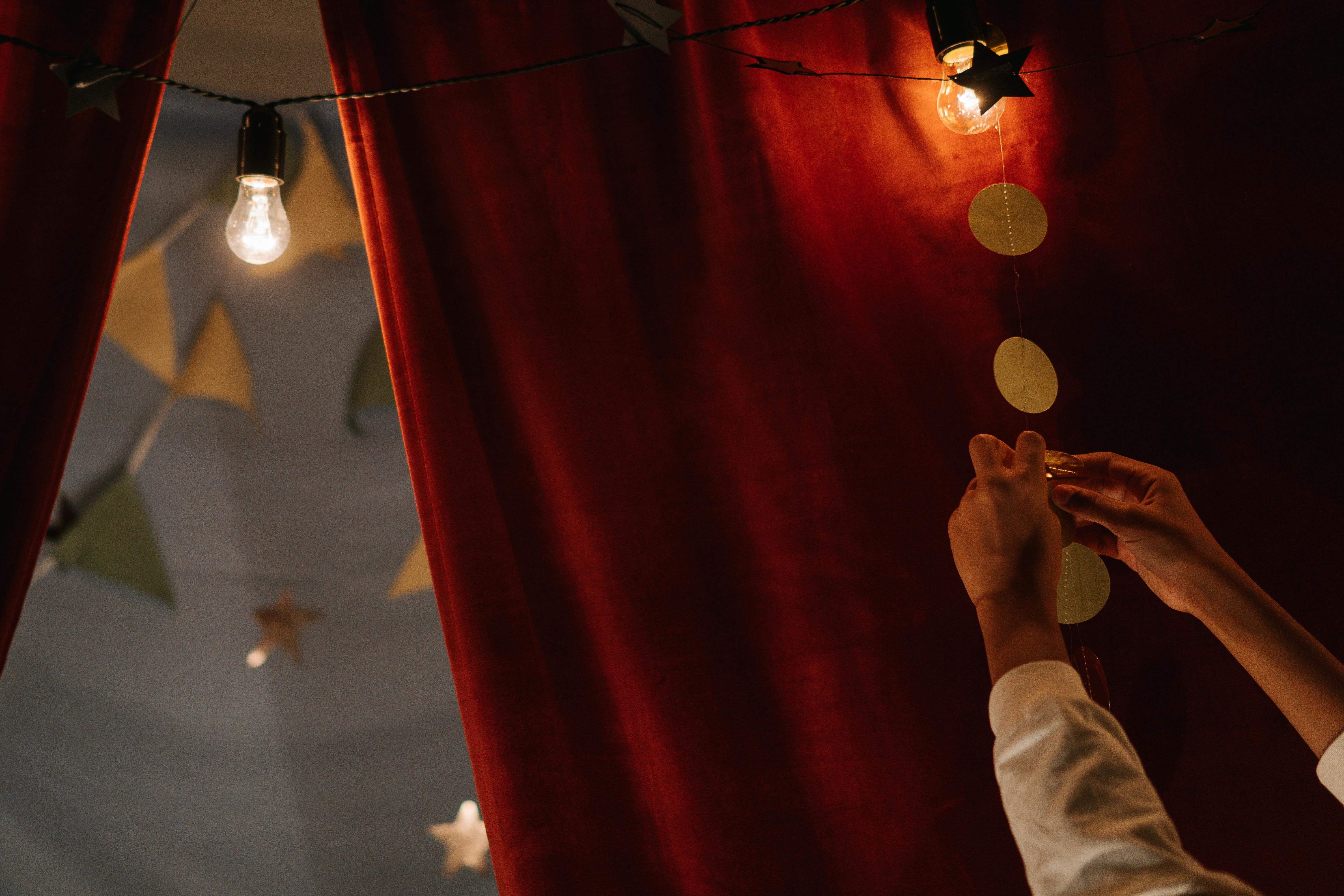 Hands decorating with ornaments against a red curtain and warm lighting.
