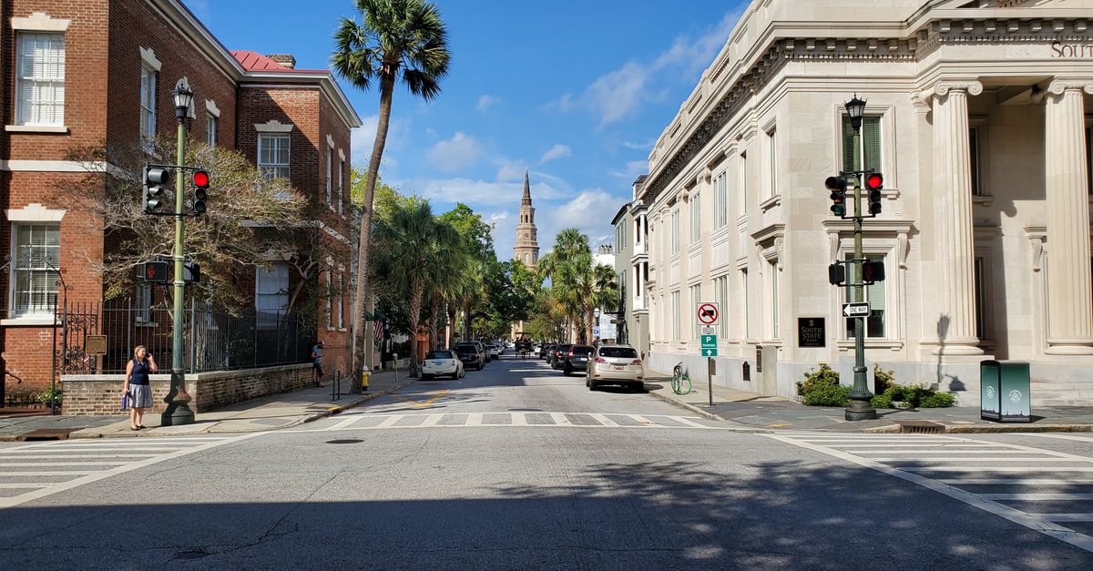View of a sunny street with historic buildings, palm trees, and a church spire in the distance.