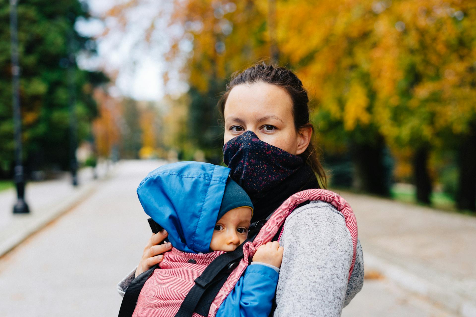 Woman wearing a mask carrying a baby in a carrier during a fall walk in the park.