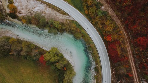 Drone view of empty narrow road near transparent river between colorful vegetation in fall countryside