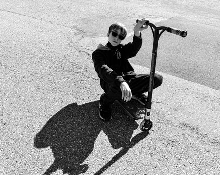 Monochrome photo of a teen sitting on a scooter, casting a strong shadow on pavement.