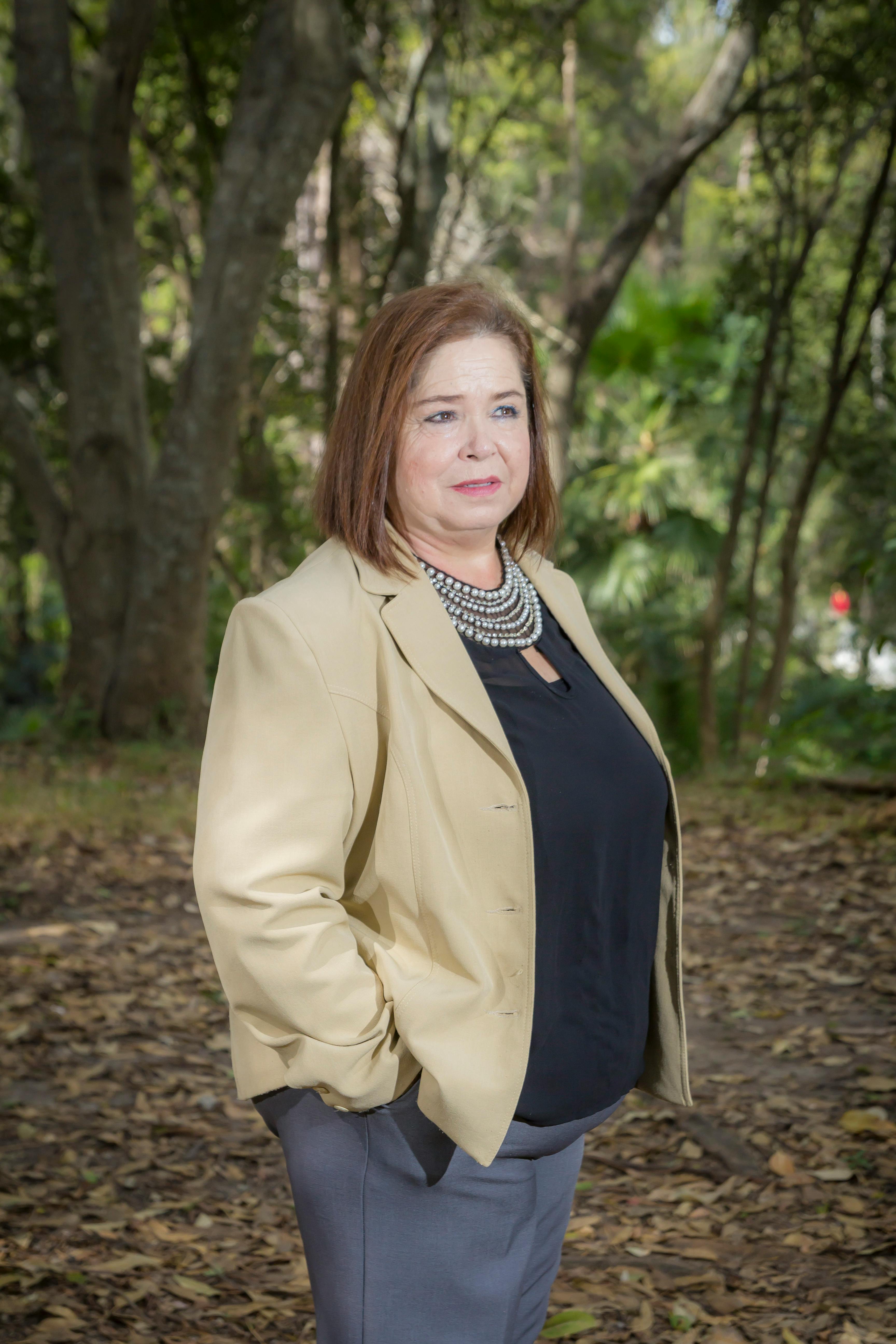 Businesswoman in a beige blazer confidently poses in a forested setting.