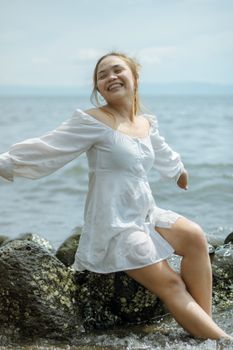 Cheerful young Asian female in white dress enjoying sun and sea water while sitting on rocks with hands sideways