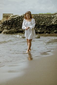 Full body of calm young barefoot ethnic female in white dress walking on wet sandy beach and looking down