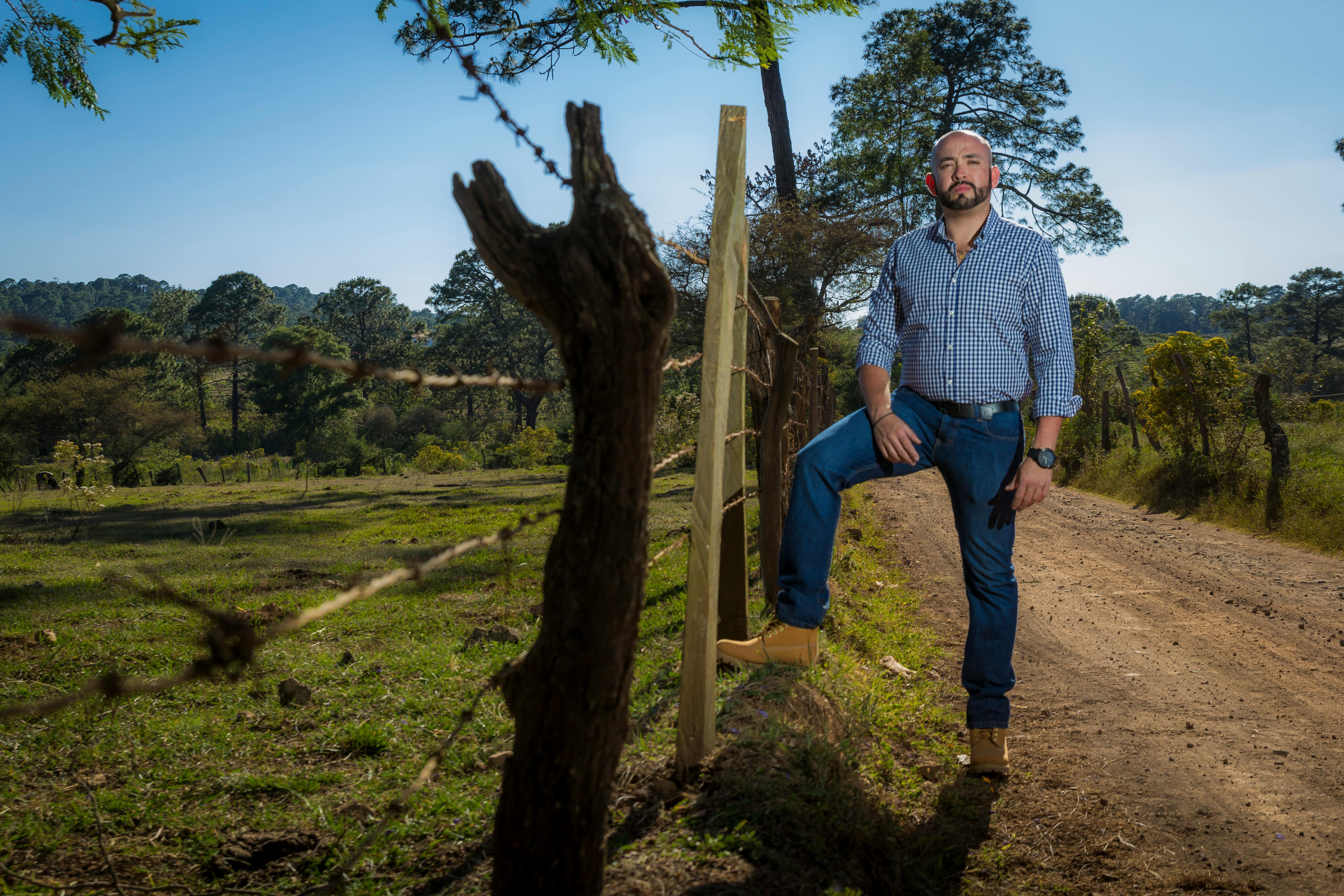 A confident man poses next to a fence in a vast rural landscape, showcasing the beauty of nature.