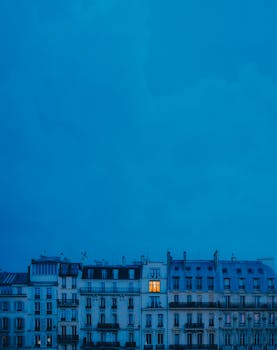 A single illuminated window stands out in an evening cityscape, creating a mood of solitude.