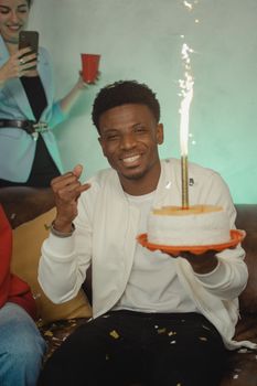 Smiling man holding a cake with a sparkler, celebrating a birthday indoors.
