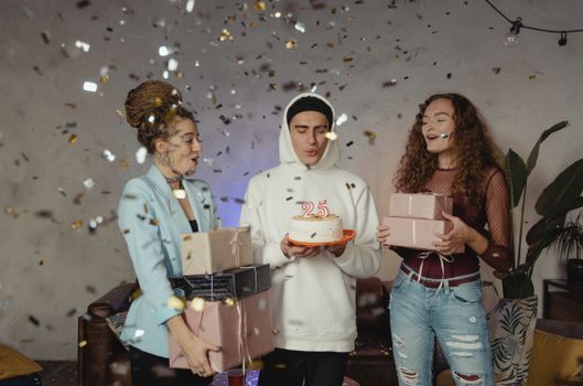Young adults celebrate a 25th birthday with cake, presents, and confetti indoors.