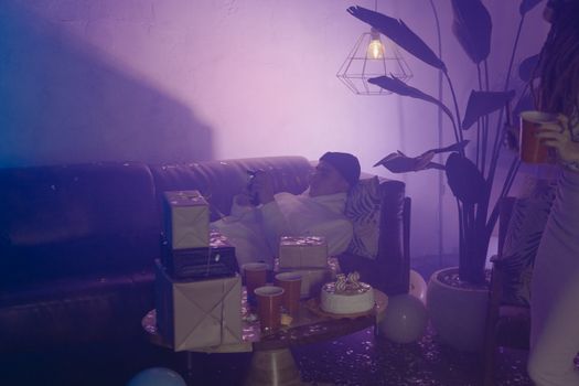 A relaxed young man laying on a couch amidst party decorations, gifts, and cake.