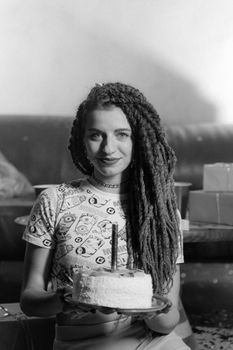Caucasian woman with dreadlocks holding a birthday cake, smiling.