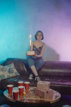 Woman holding a sparkler cake on a couch, surrounded by birthday decorations.