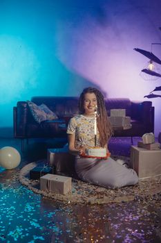 Woman sitting in a confetti-filled room holding a birthday cake with sparklers, surrounded by gifts.