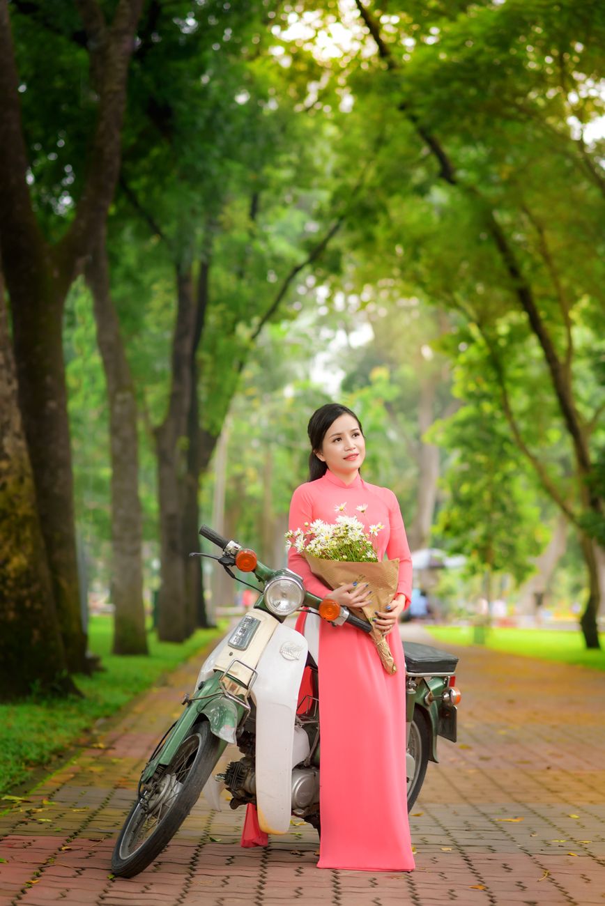 Asian woman wearing ao dai holding flowers, standing by a scooter in a park.