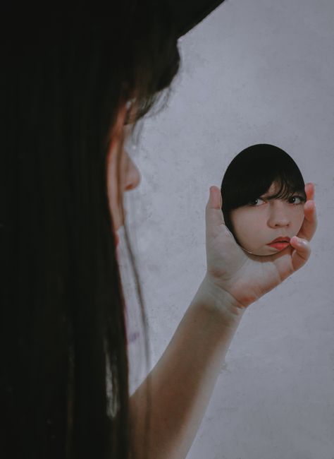 Crop serious ethnic female with long hair and red lips reflecting in small hand mirror