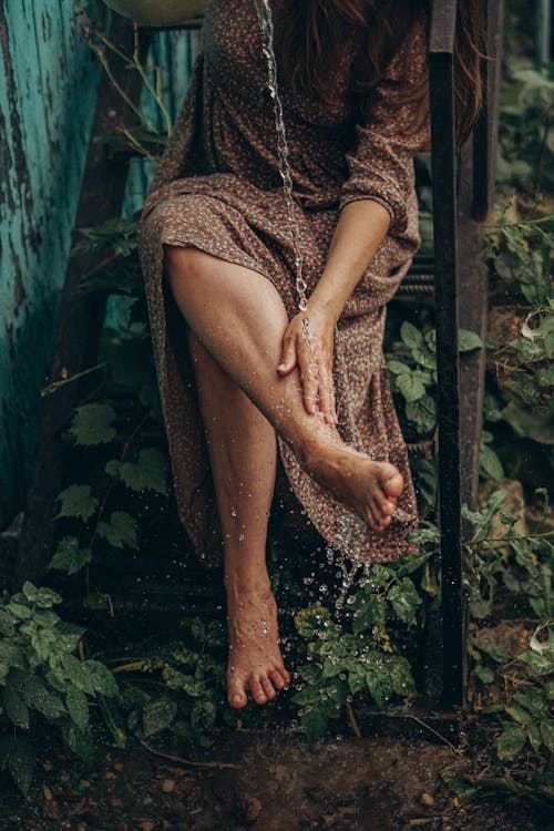 Crop faceless barefoot female washing legs with clear water while sitting on metal staircase overgrown with greenery