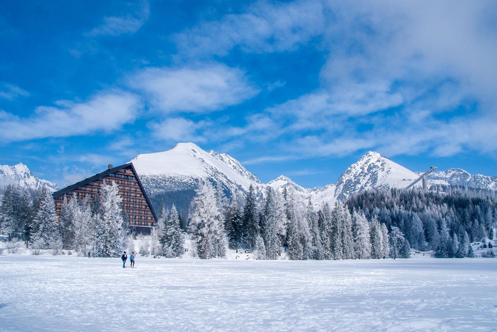 Two people walking in a snowy landscape near a winter resort and snowy mountains.