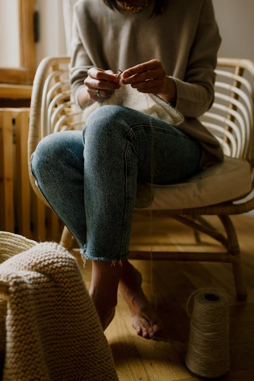 Crop unrecognizable barefoot woman in jeans and warm sweater knitting warm cloth with spool of thread and needles while sitting alone at home