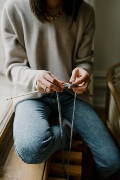 A woman skillfully knitting with needles, sitting by a window indoors, wearing casual attire.
