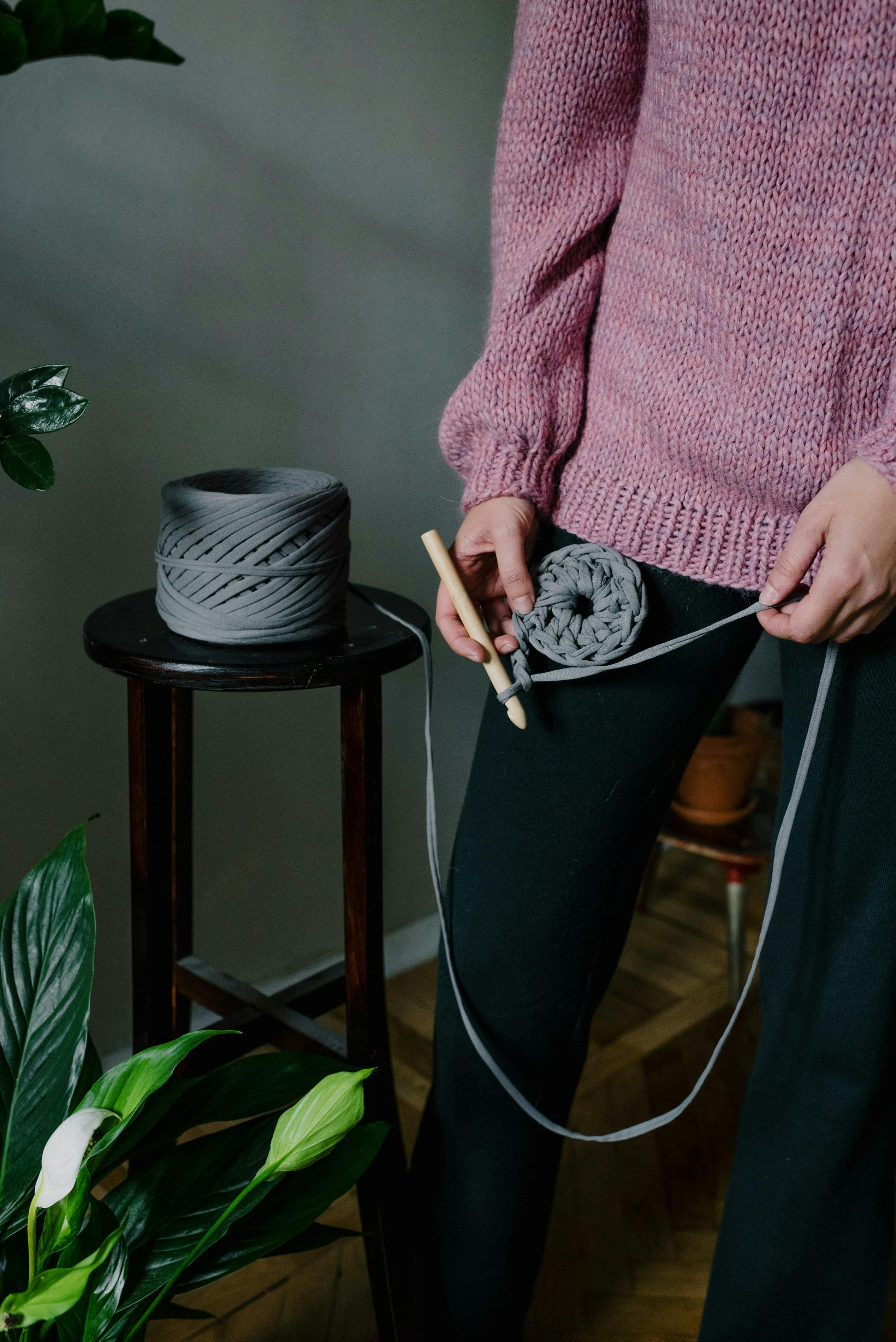 Close-up of a woman crocheting a pink sweater with chunky yarn in an indoor setting.