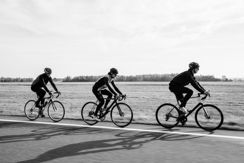 Free Three cyclists biking on a road through a flat countryside landscape in black and white. Stock Photo