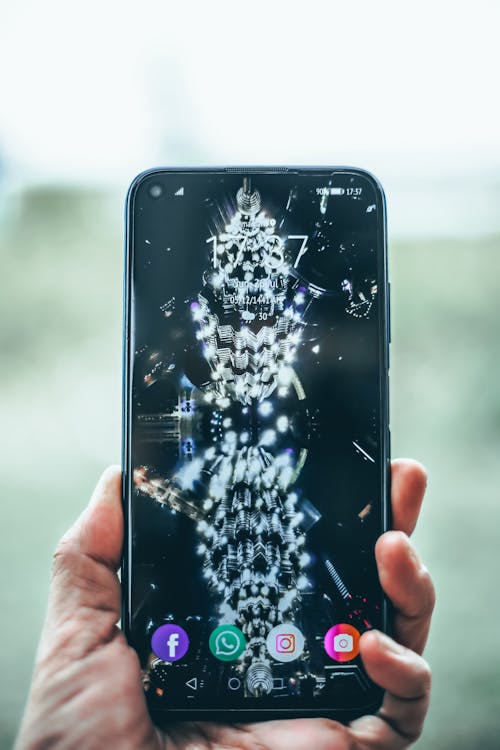 Crop person hand holding modern mobile phone with various apps and illuminated city on wallpaper