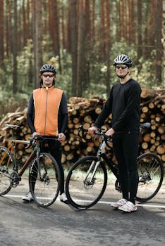 Two men with bicycles standing in front of wood logs in a forest setting, wearing helmets and activewear.