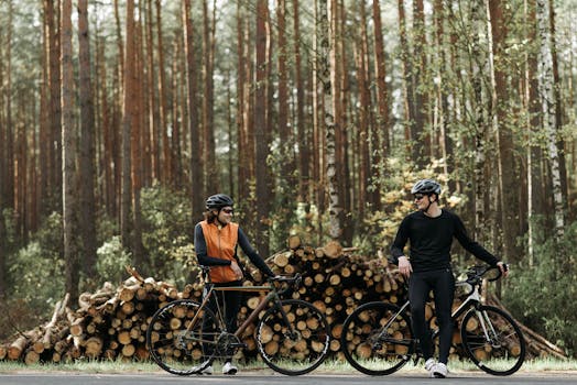 Two cyclists in sportswear pause by stacked logs in a serene forest backdrop.