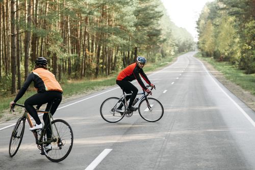 Free Two cyclists ride on an open road surrounded by lush greenery, enjoying a sunny day outdoors. Stock Photo